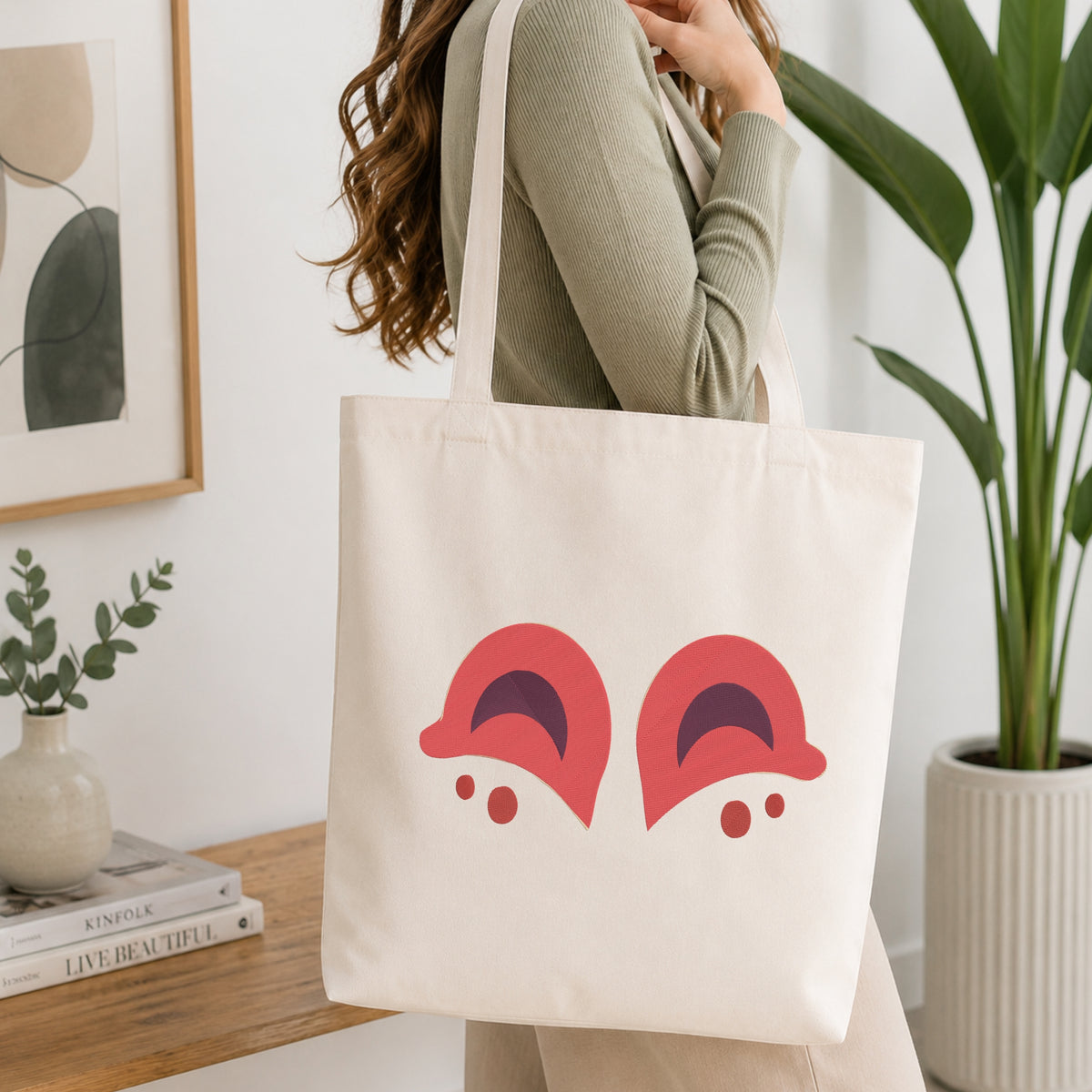 A person wearing a sage green ribbed long-sleeve top carries a cream canvas tote bag with a graphic of two red cartoon eyes on the front, set in a bright, modern room with a wooden table, framed art, and a potted plant.