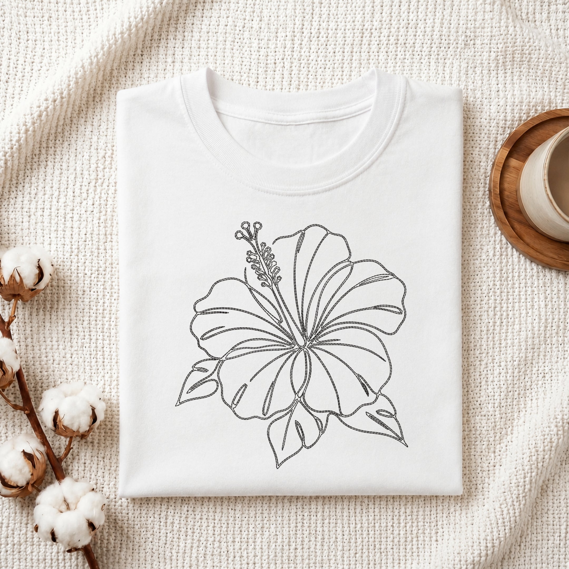 A folded white crewneck t-shirt with a black line art drawing of a hibiscus flower, styled on a textured cream knit blanket with cotton bolls and a wooden cup in the background.