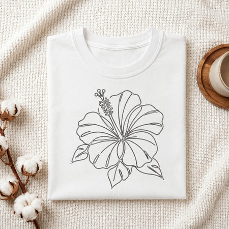 A folded white crewneck t-shirt with a black line art drawing of a hibiscus flower, styled on a textured cream knit blanket with cotton bolls and a wooden cup in the background.