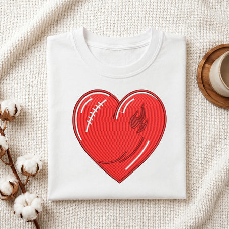 A folded white crewneck t-shirt with a red heart-shaped graphic on the front, styled in a flat-lay arrangement on a textured cream fabric with cotton bolls and a wooden cup.