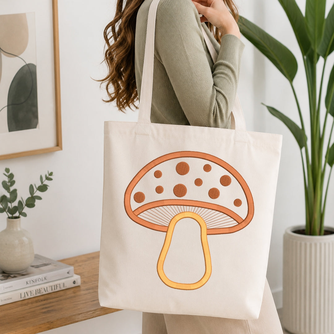 A woman wearing a sage green ribbed sweater carries a cream canvas tote bag with a large orange and yellow mushroom graphic printed on the front, set in a bright, modern room with a wooden table, books, and a potted plant.