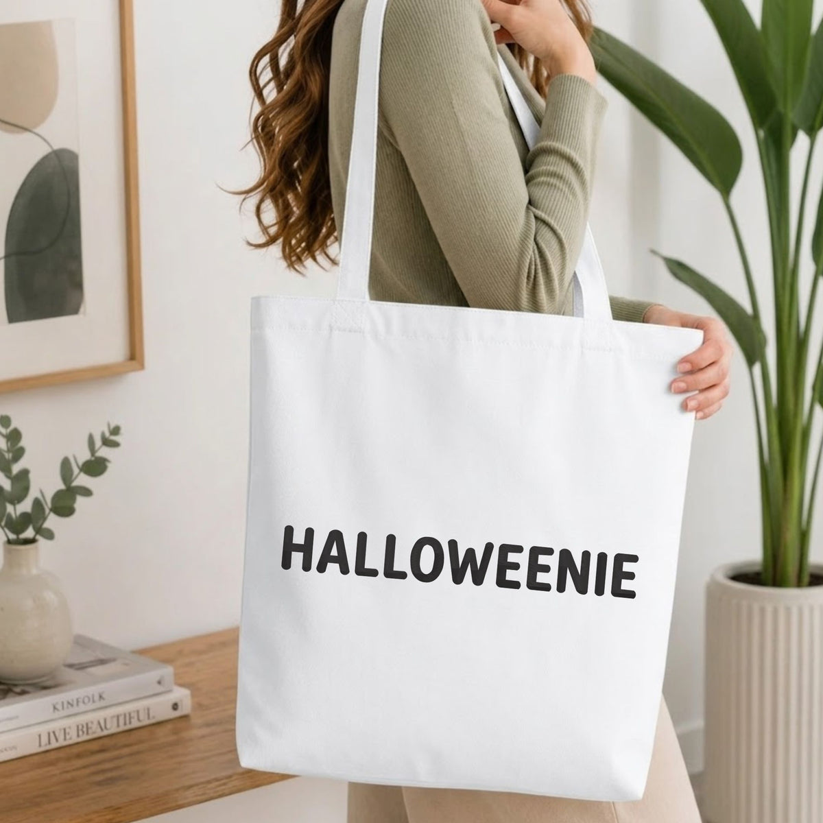 Woman carrying a white natural canvas tote bag featuring a "HALLOWEENIE" machine embroidery design in a bright, modern interior setting   
