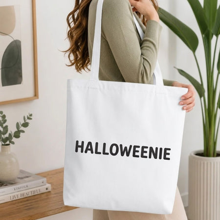 Woman carrying a white natural canvas tote bag featuring a "HALLOWEENIE" machine embroidery design in a bright, modern interior setting   