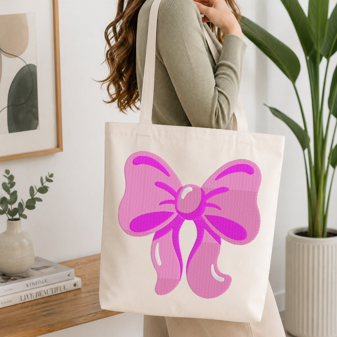 A woman wearing a sage green ribbed sweater carries a cream canvas tote bag with a large pink bow graphic on the front, set in a bright, modern room with a wooden table and potted plants.