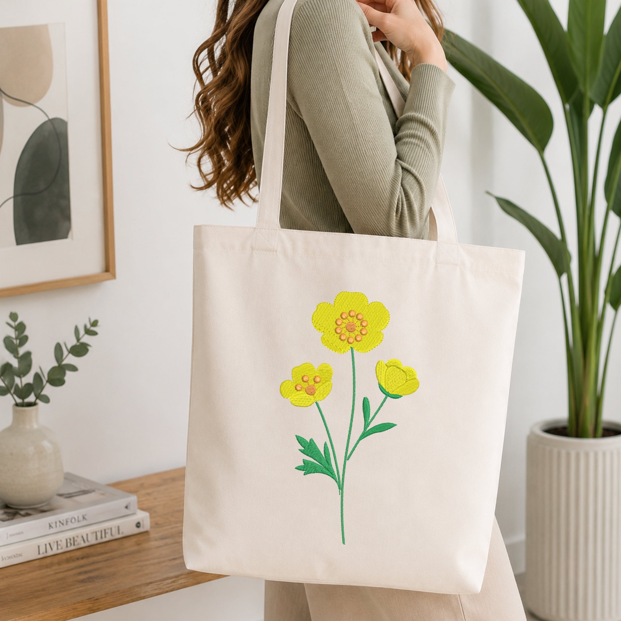 A woman wearing a sage green ribbed long-sleeve top carries a cream canvas tote bag with a yellow flower and green leaves graphic printed on the front, set in a bright, modern room with a wooden table and potted plants.