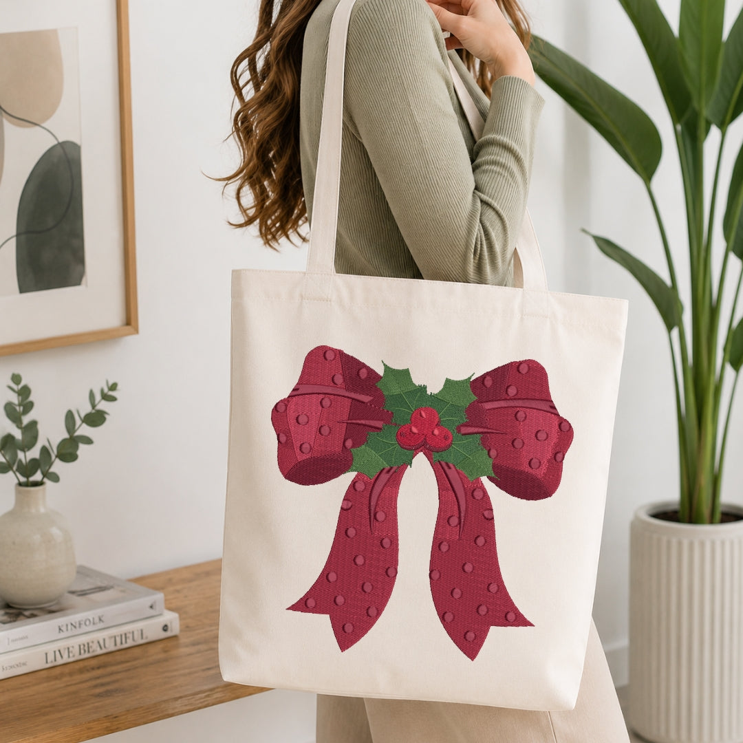 A woman wearing a sage green ribbed sweater carries a cream canvas tote bag decorated with a large red bow and green holly leaves on the front, set in a bright, modern room with a wooden table and potted plants.
