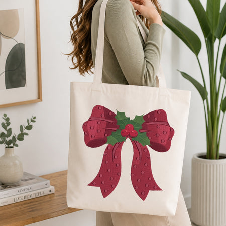 A woman wearing a sage green ribbed sweater carries a cream canvas tote bag decorated with a large red bow and green holly leaves on the front, set in a bright, modern room with a wooden table and potted plants.