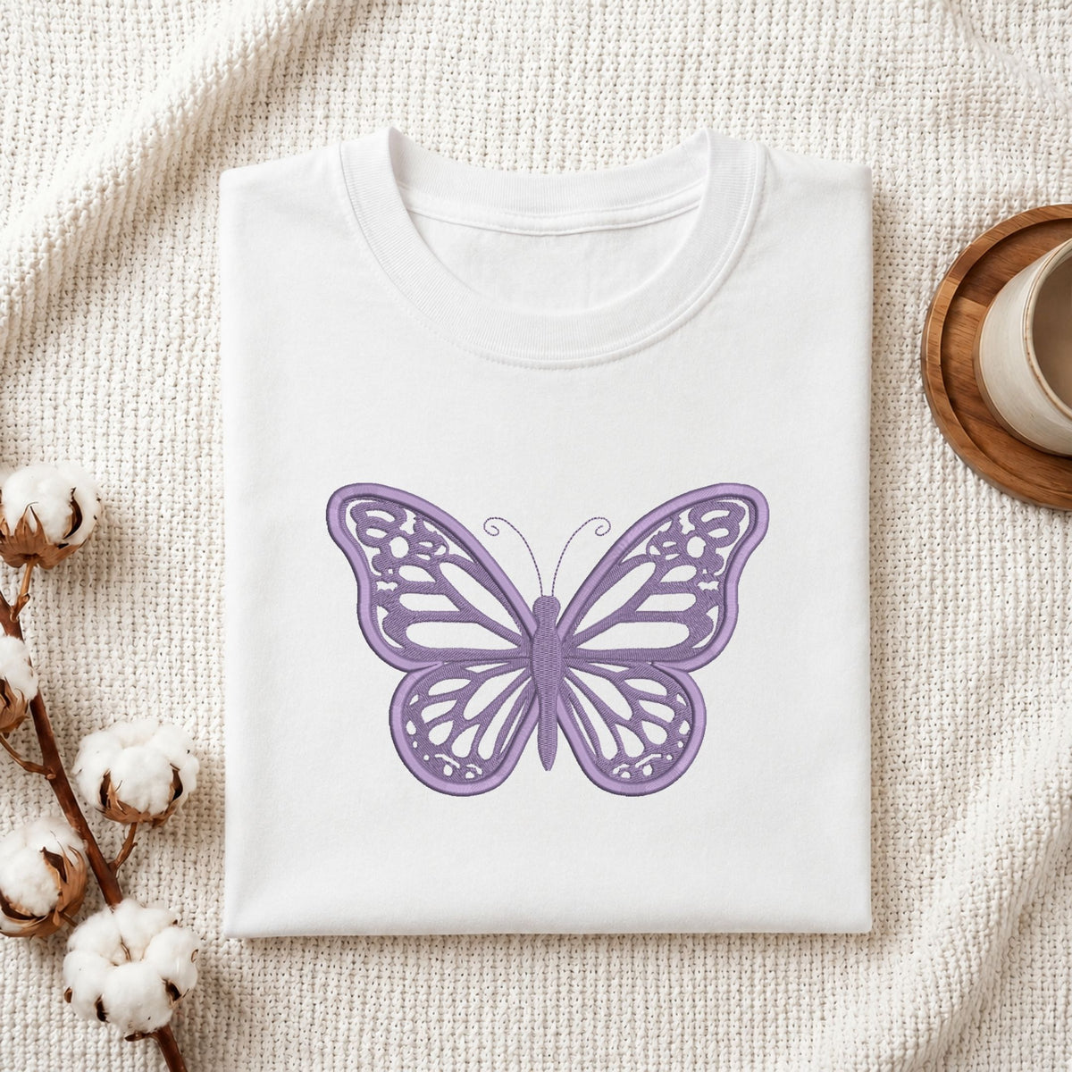 Folded white cotton t-shirt mockup featuring a purple butterfly machine embroidery design, styled with cotton stems and a wooden cup