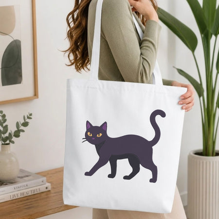 A woman wearing a sage green ribbed sweater carries a white canvas tote bag featuring a stylized black cat illustration with yellow eyes, set against a bright, modern indoor background with a wooden table and potted plants.
