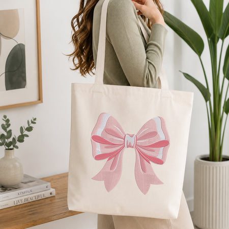 An off-white canvas tote bag with a large pink bow graphic on the front, carried over the shoulder of a woman in a sage green ribbed top. The scene is set in a bright, modern room with a wooden table, books, a framed print, and a potted plant.