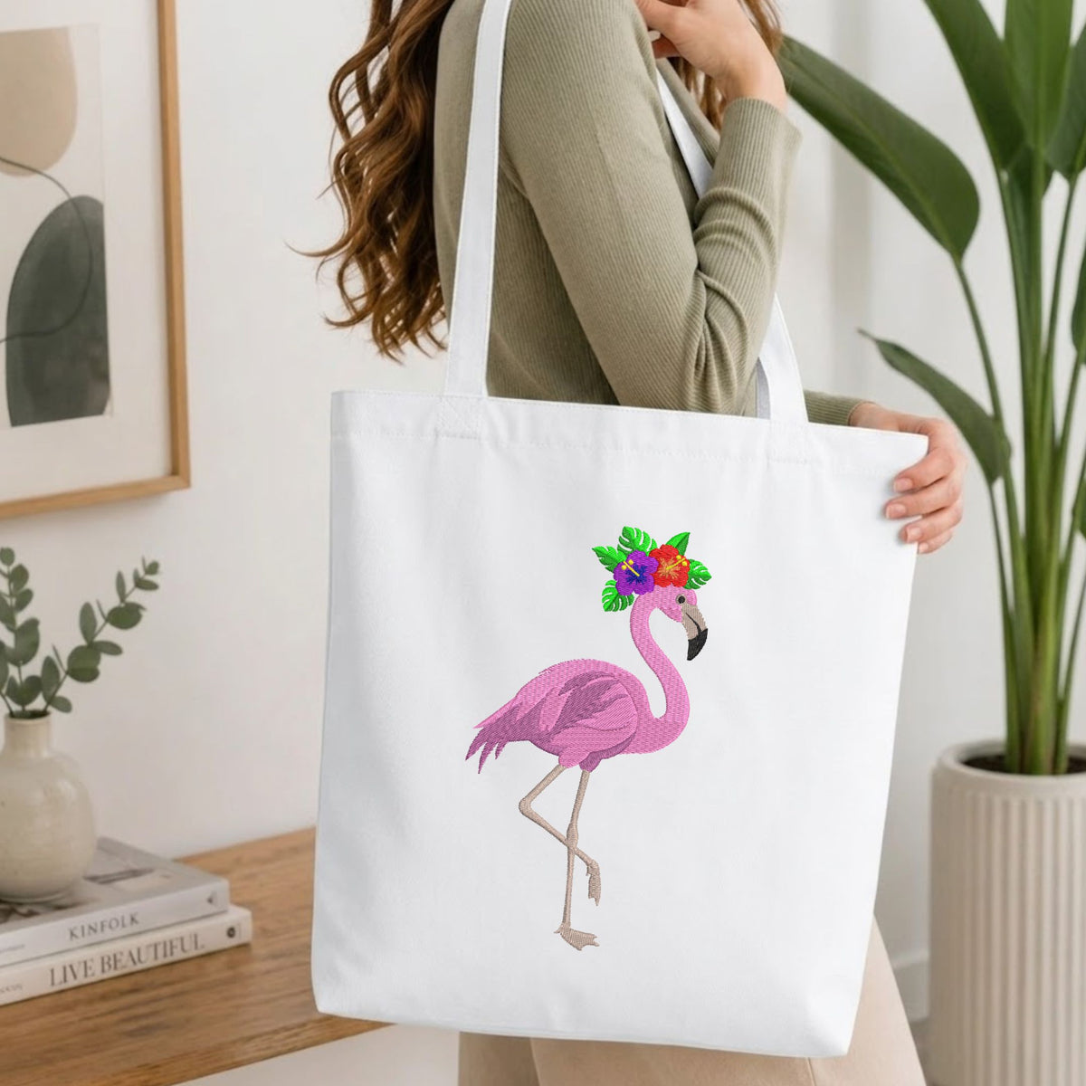 A woman wearing a sage green ribbed sweater carries a white canvas tote bag. The bag features a graphic of a pink flamingo wearing a colorful green and orange flower crown.
