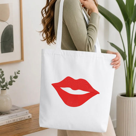 Woman carrying a white natural canvas tote bag featuring an embroidered red lips design in a bright, modern interior setting