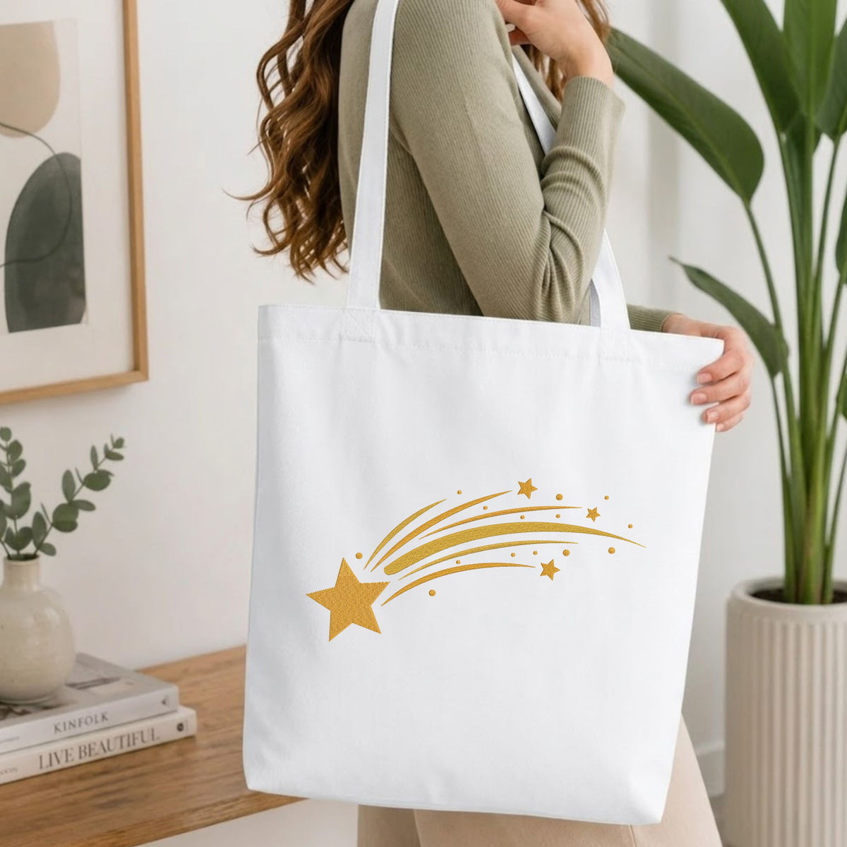 Woman carrying a white natural canvas tote bag featuring an embroidered golden shooting star design in a bright, modern interior setting