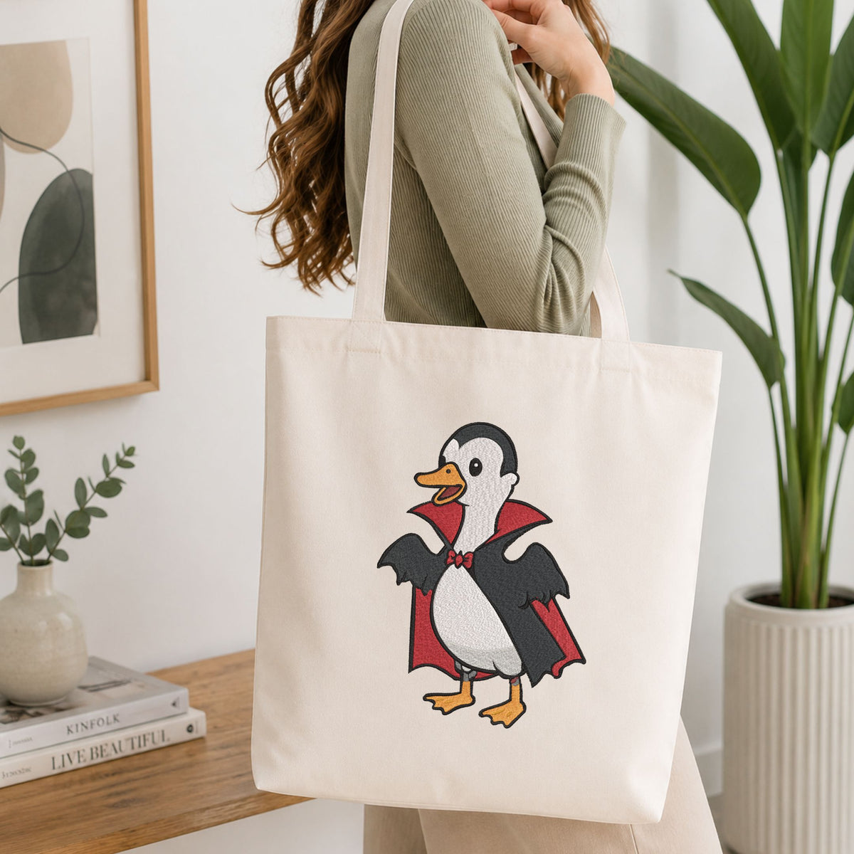 Woman carrying a white natural canvas tote bag featuring a cute duck in a vampire Dracula costume Halloween machine embroidery design in a bright, modern interior setting