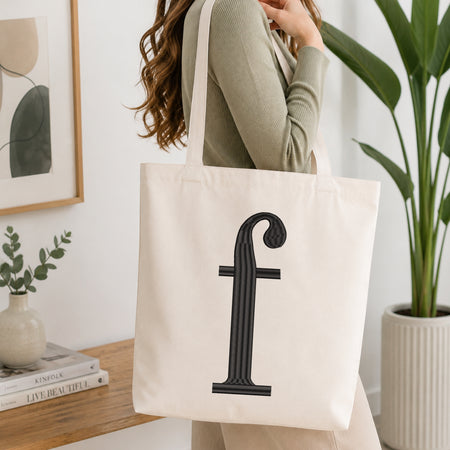 A woman wearing a sage green ribbed sweater carries a large off-white canvas tote bag with a black serif letter 'f' printed on the front, set in a bright, modern room with a wooden table, books, and a potted plant.