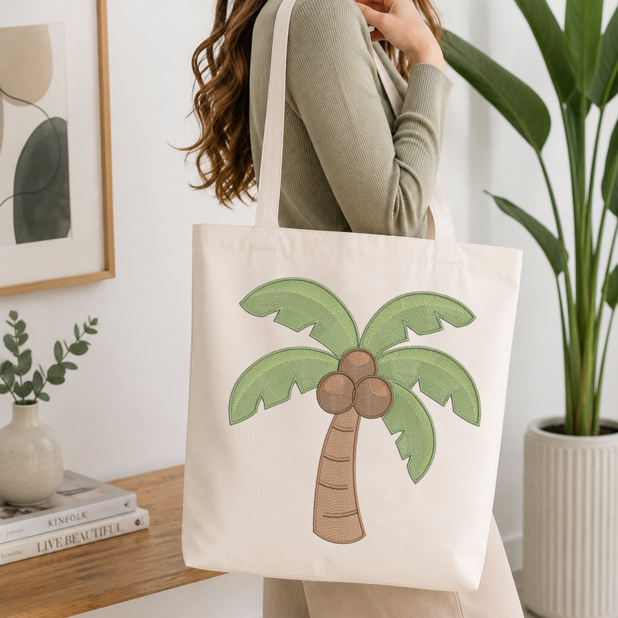 A woman wearing a sage green ribbed long-sleeve top carries a cream canvas tote bag with a green palm tree and brown coconuts printed on the front, set in a bright, modern room with a wooden table, framed art, and a potted plant.