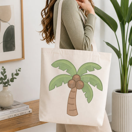 A woman wearing a sage green ribbed long-sleeve top carries a cream canvas tote bag with a green palm tree and brown coconuts printed on the front, set in a bright, modern room with a wooden table, framed art, and a potted plant.