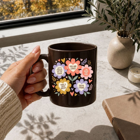 A hand holds a dark brown ceramic mug decorated with colorful floral illustrations and motivational quotes, set on a light stone tabletop near a window with a potted plant and candle in the background.