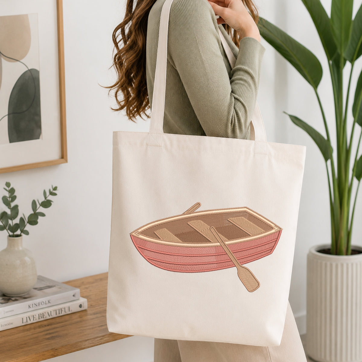 A woman wearing a sage green ribbed long-sleeve top carries a cream canvas tote bag with an illustration of a pinkish-red rowboat and wooden oars. The scene is set in a bright, modern room with a wooden table, books, a small plant in a vase, and a large green potted plant in the background.