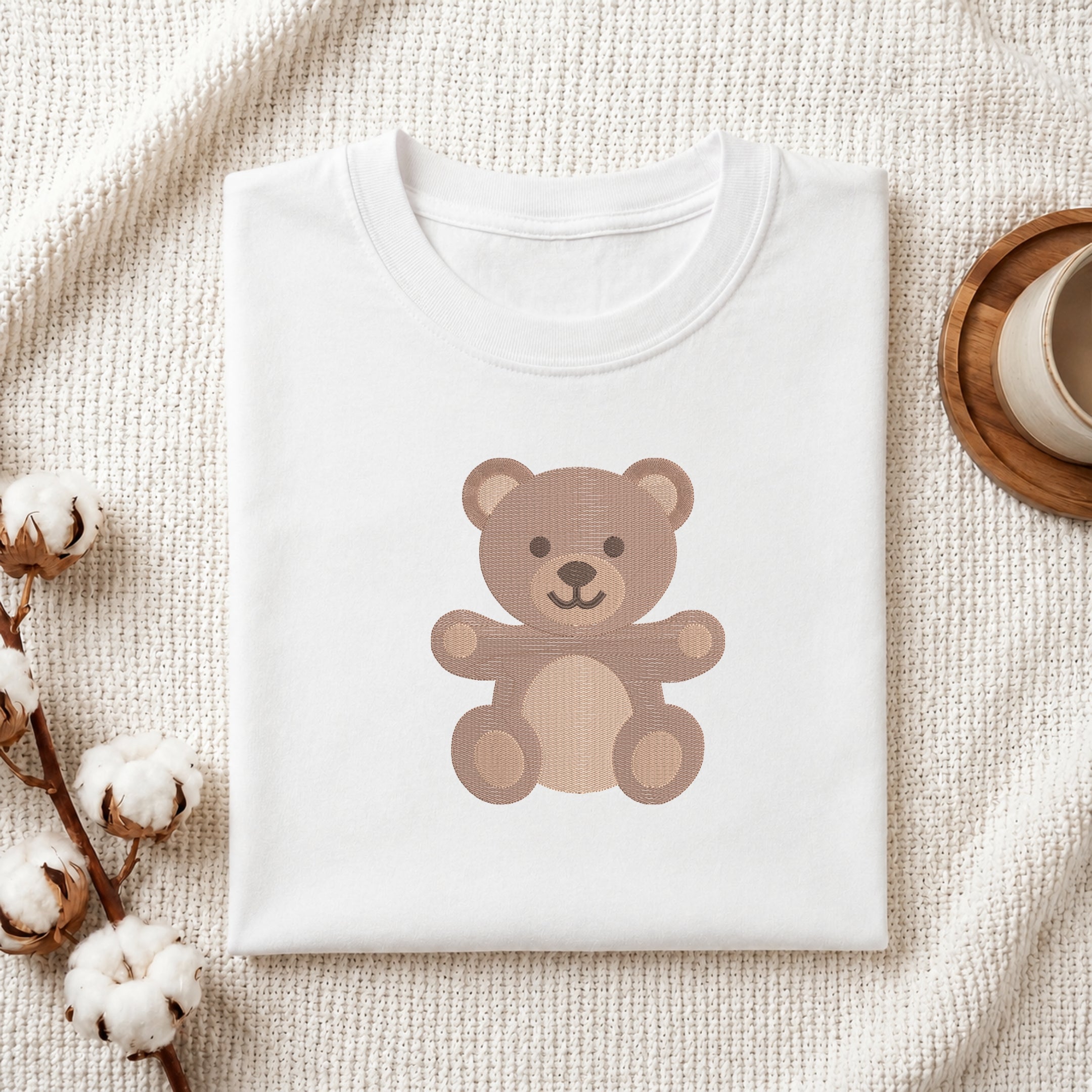 A folded white T-shirt with a brown teddy bear graphic on the front, styled on a textured cream fabric with cotton bolls and a wooden cup edge visible.