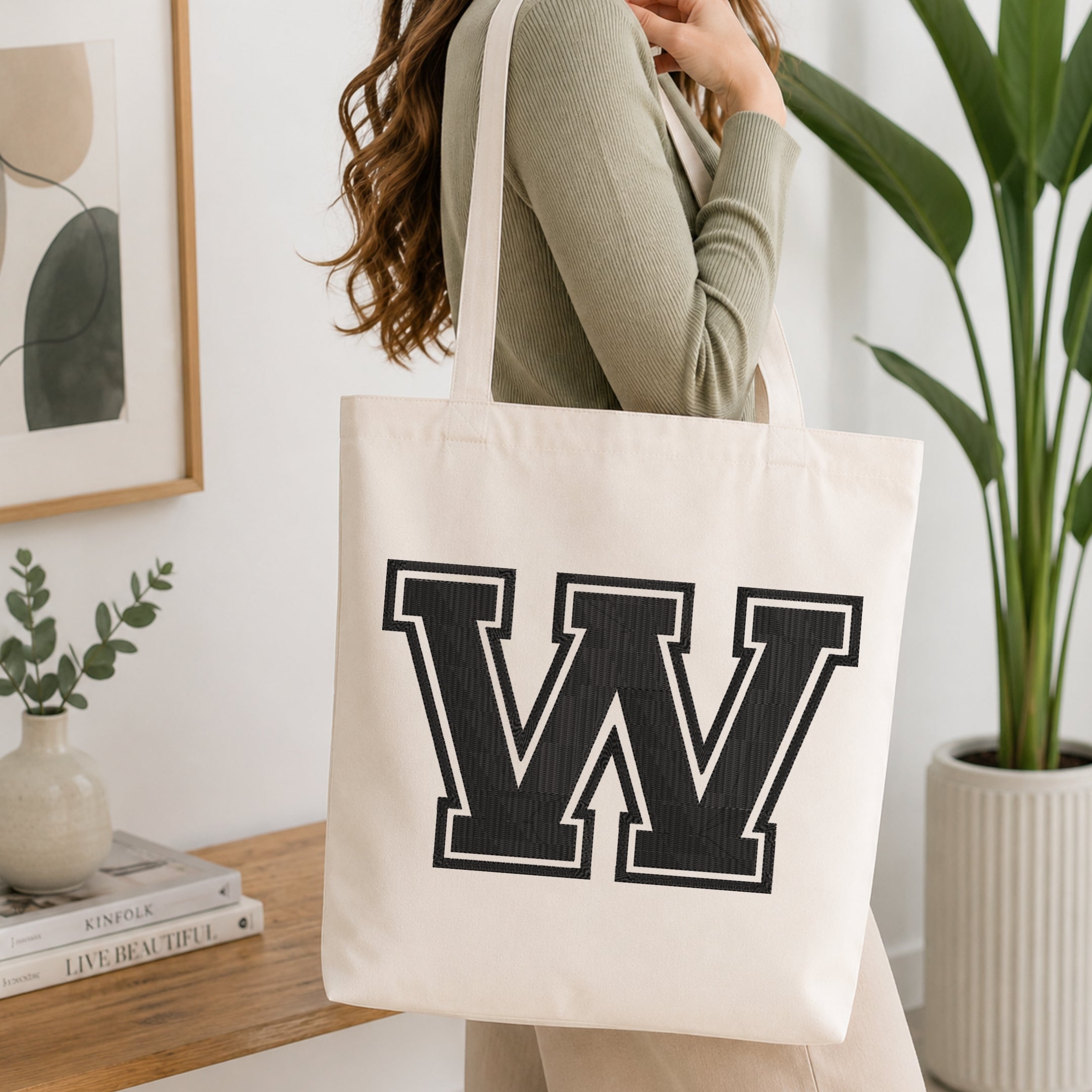 A woman wearing a sage green ribbed long-sleeve top carries a large off-white canvas tote bag with a bold black block letter 'W' outlined in white on the front. The scene is set in a bright, modern room with a wooden console table, a framed print, a small vase with greenery, and a potted plant.