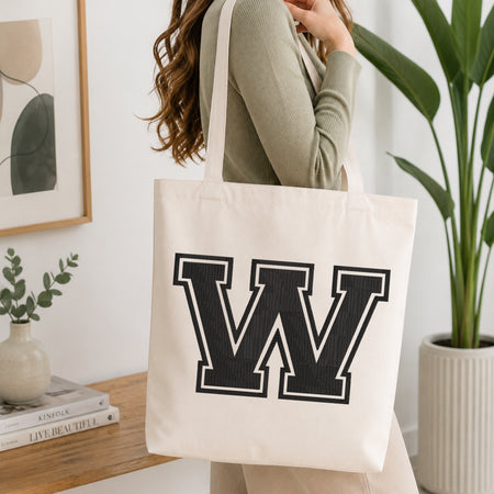 A woman wearing a sage green ribbed long-sleeve top carries a large off-white canvas tote bag with a bold black block letter 'W' outlined in white on the front. The scene is set in a bright, modern room with a wooden console table, a framed print, a small vase with greenery, and a potted plant.