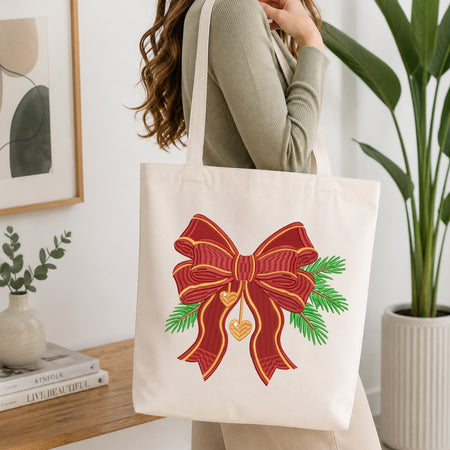 An off-white canvas tote bag with a large red embroidered bow, green pine branches, and a gold heart, carried on the shoulder of a woman in a cozy indoor setting.