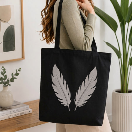 A woman wearing a beige ribbed sweater carries a black canvas tote bag with two large white feather designs on the front, set in a bright, modern room with a potted plant and books on a wooden table.