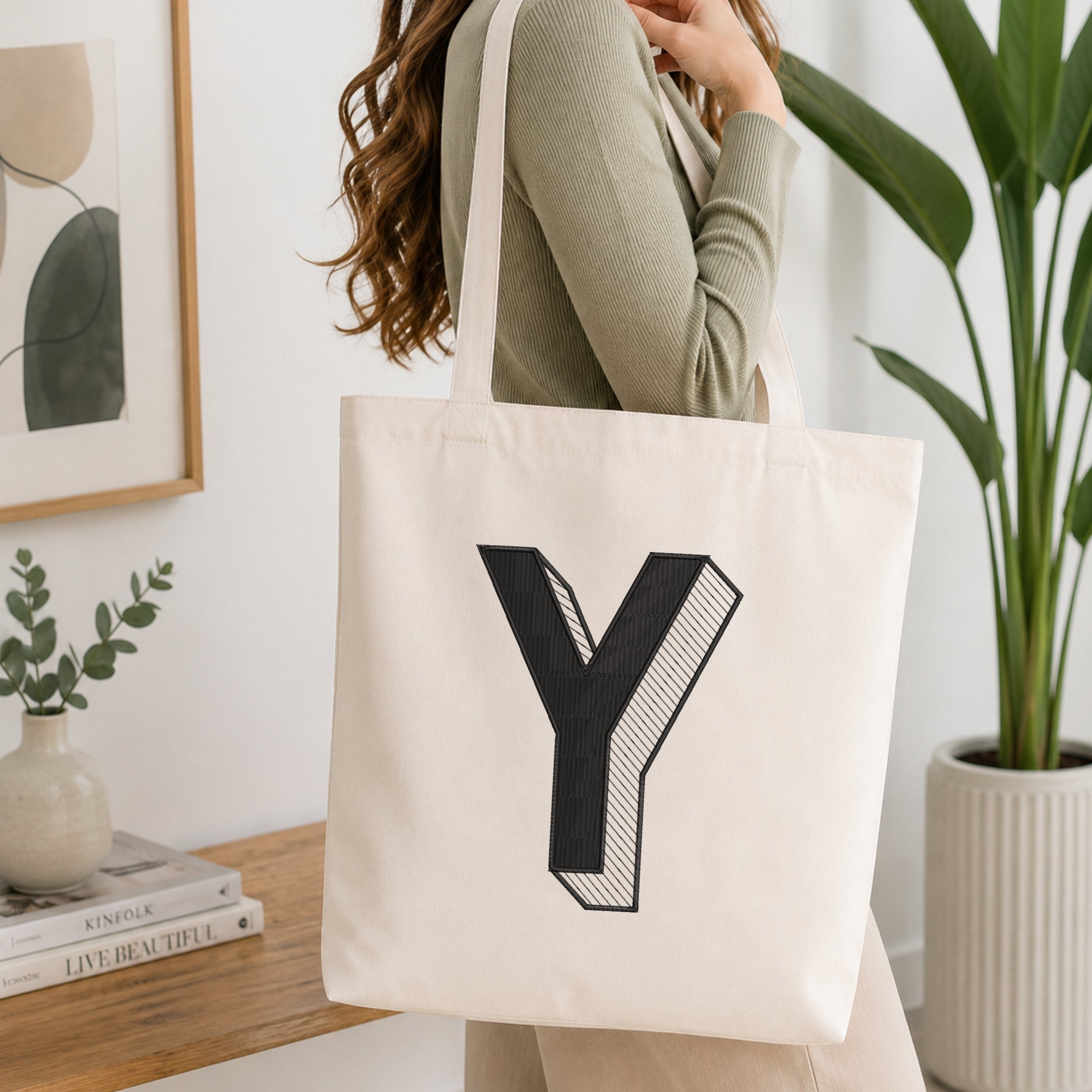 A woman wearing a sage green ribbed long-sleeve top carries a cream canvas tote bag with a large black letter 'Y' printed on the front, set in a bright, modern room with a wooden table, books, and a potted plant.