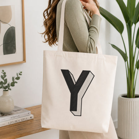 A woman wearing a sage green ribbed long-sleeve top carries a cream canvas tote bag with a large black letter 'Y' printed on the front, set in a bright, modern room with a wooden table, books, and a potted plant.