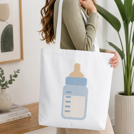 A woman in a sage green ribbed top carries a white canvas tote bag featuring a stylized baby bottle graphic in light blue, beige, and white. The setting is a bright, modern room with a wooden table, books, a framed print, and a potted plant.