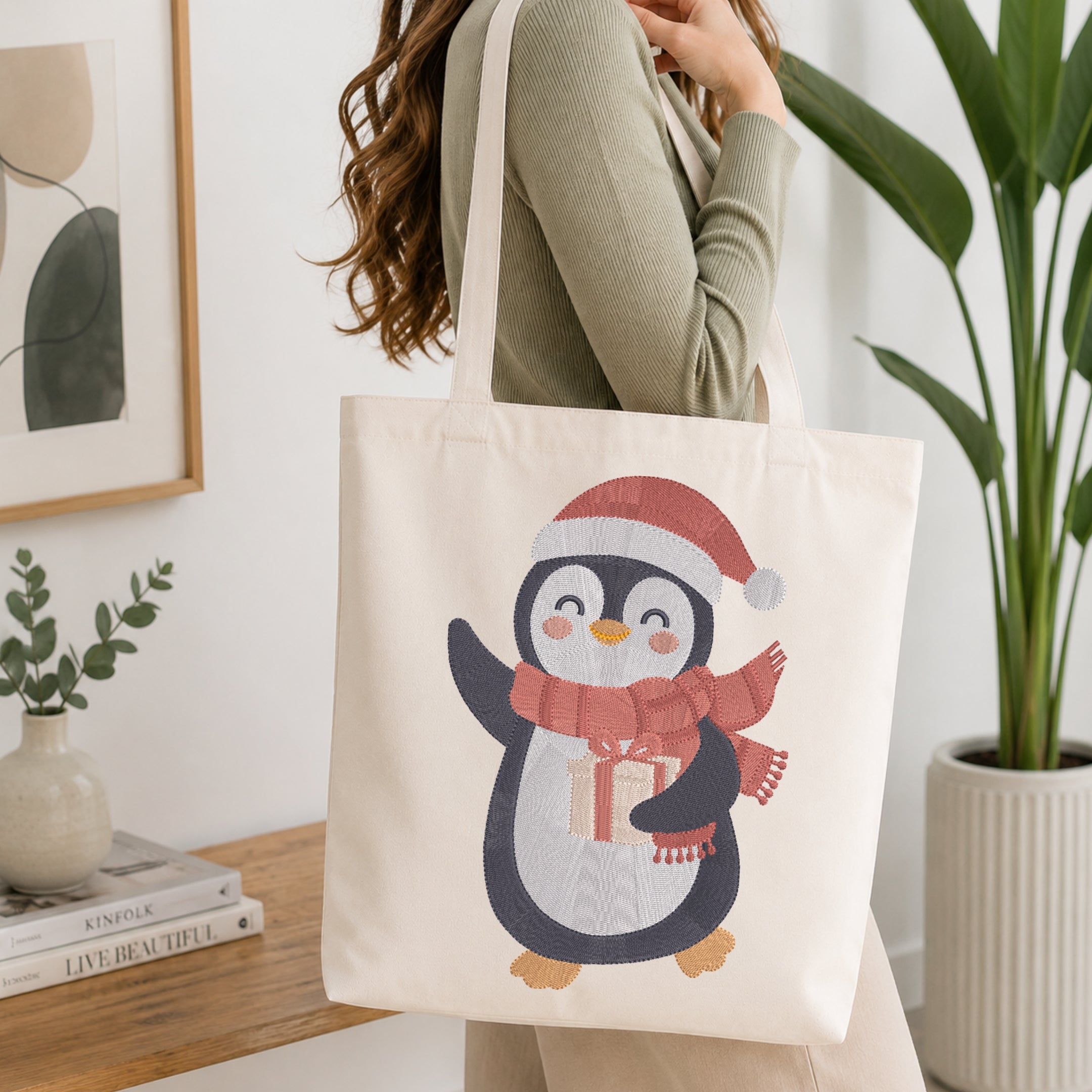 A woman wearing a sage green ribbed sweater carries a cream canvas tote bag featuring a cute penguin illustration wearing a red Santa hat and scarf with a gift box.