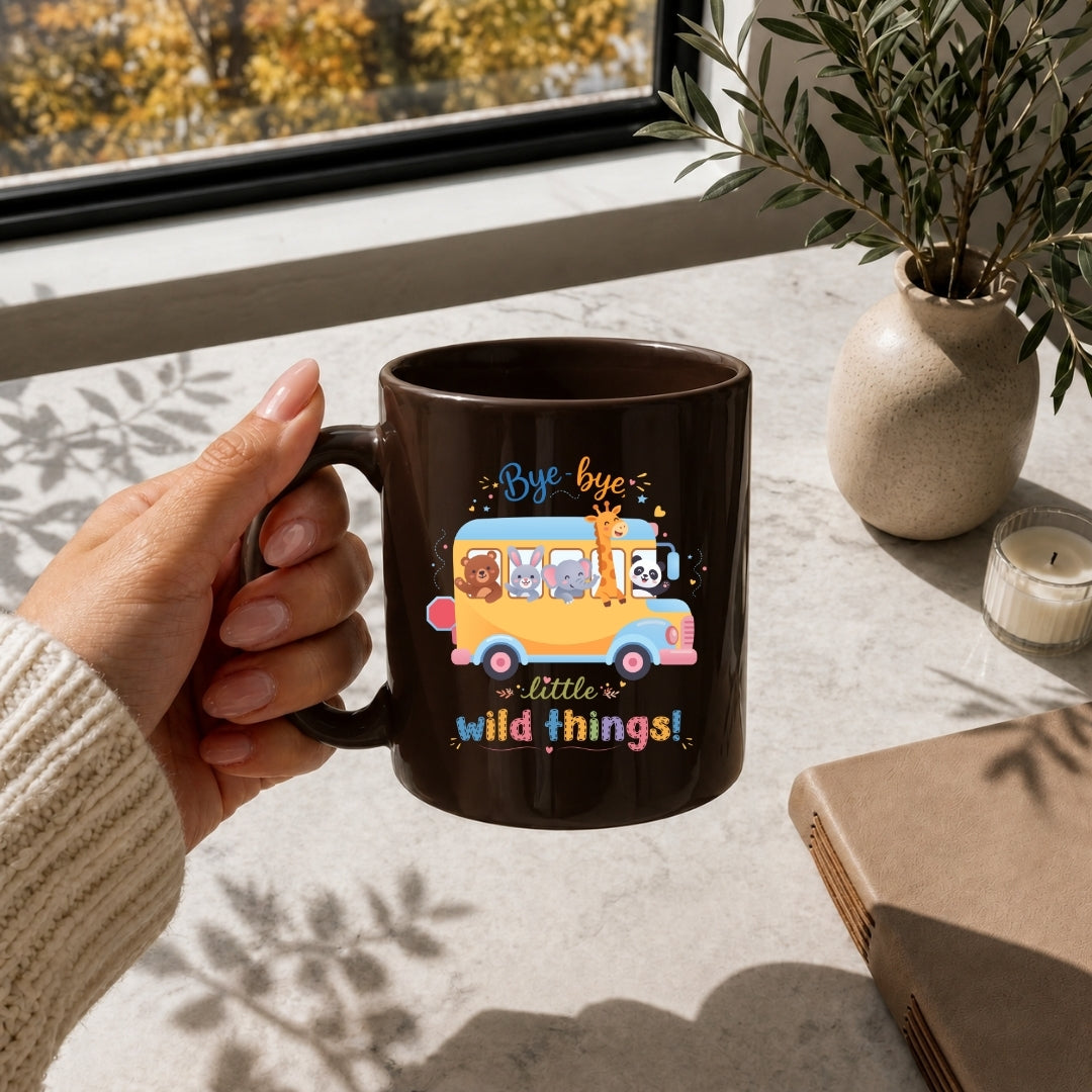 A hand holding a brown ceramic mug with a school bus design featuring cartoon animals and the text "Bye-bye little wild things!"