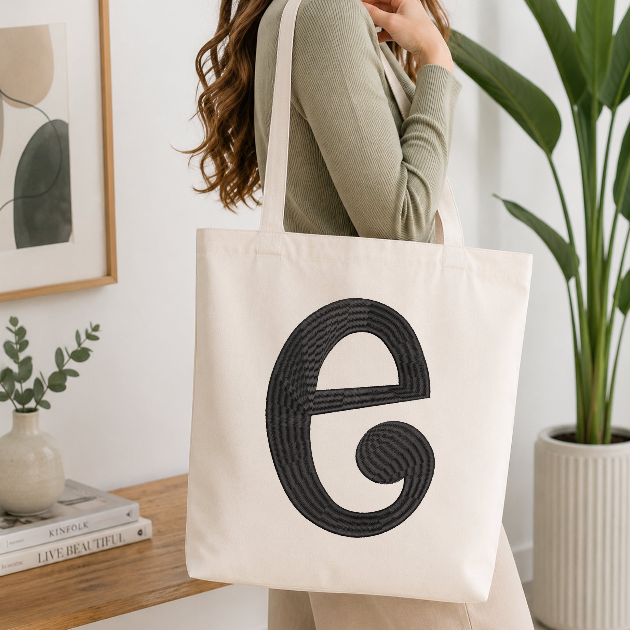 A woman wearing a sage green ribbed long-sleeve top carries a large off-white canvas tote bag with a bold black spiral 'e' monogram on the front, set in a bright, modern room with a wooden console table, framed art, and a potted plant.