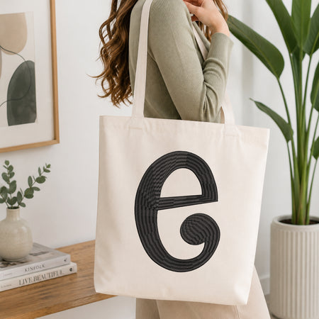 A woman wearing a sage green ribbed long-sleeve top carries a large off-white canvas tote bag with a bold black spiral 'e' monogram on the front, set in a bright, modern room with a wooden console table, framed art, and a potted plant.