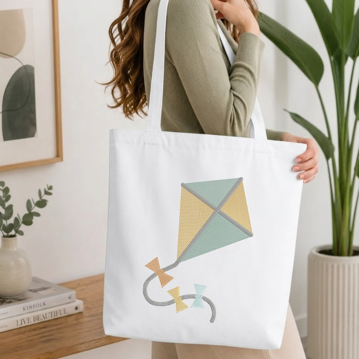 A person holding a natural canvas tote bag printed with an embroidered cute kite design, set against a bright interior room background with green plants.
