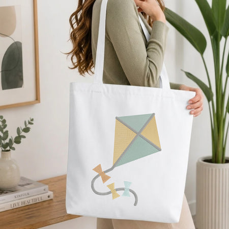 A person holding a natural canvas tote bag printed with an embroidered cute kite design, set against a bright interior room background with green plants.