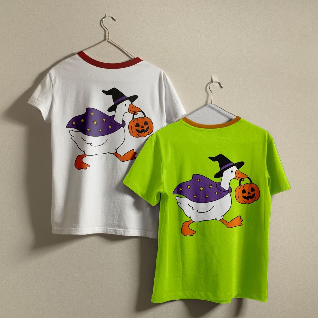 Two children's short-sleeve ringer T-shirts hanging on hangers against a plain beige wall. The left shirt is white with a red collar and shows a Halloween goose graphic on the back.