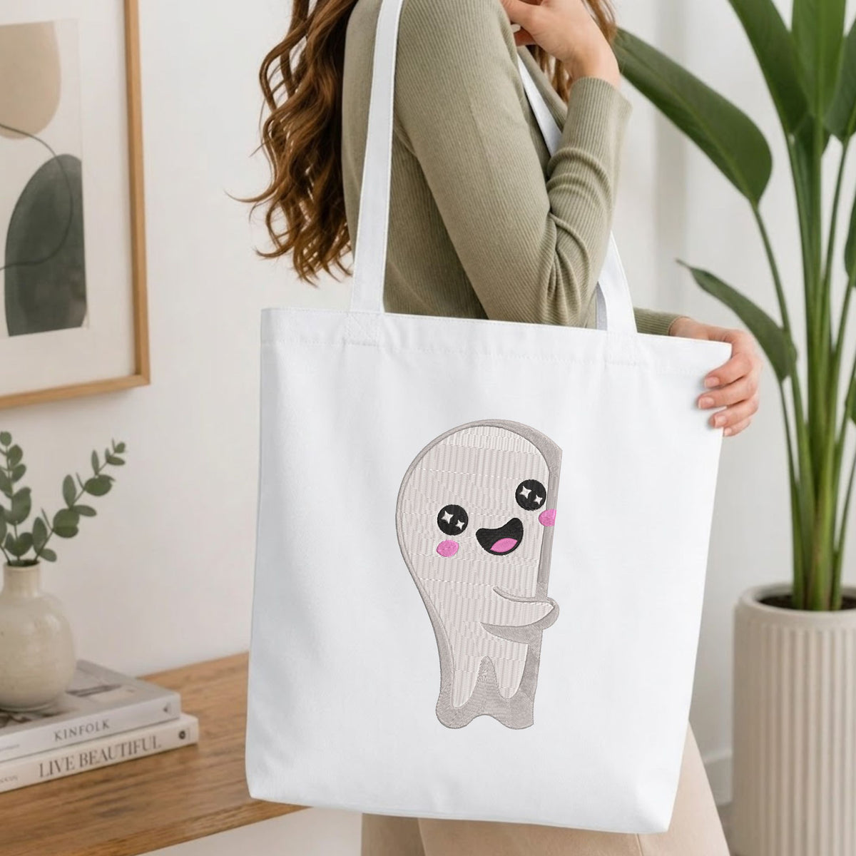 Woman carrying a white natural canvas tote bag featuring an embroidered cute ghost design in a bright, modern interior setting.