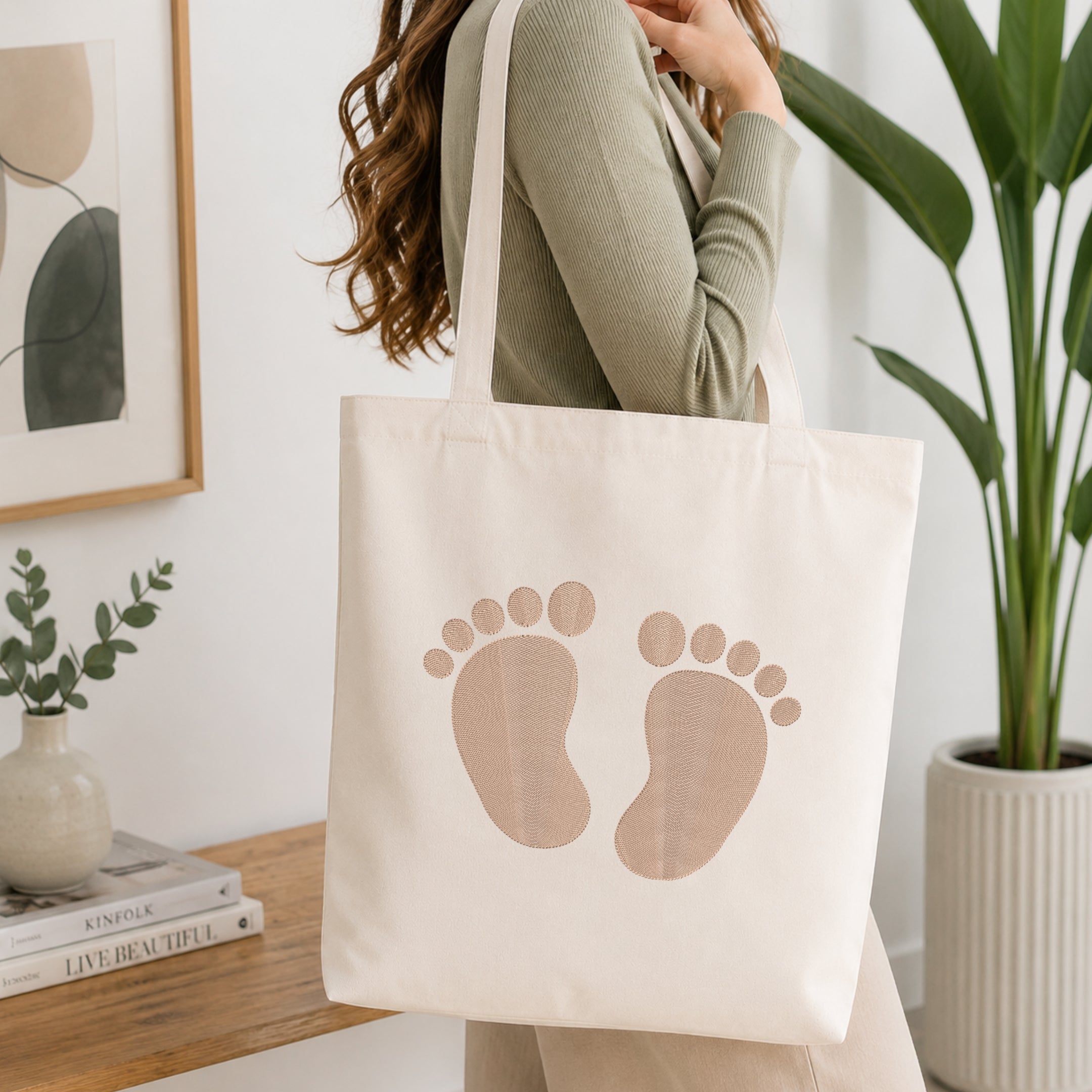 A woman wearing a sage green ribbed long-sleeve top carries a cream canvas tote bag with a printed pair of baby footprints in a warm beige color. The scene is set in a bright, modern room with a wooden console table, a framed print, a small potted plant, and a larger green houseplant in the background.