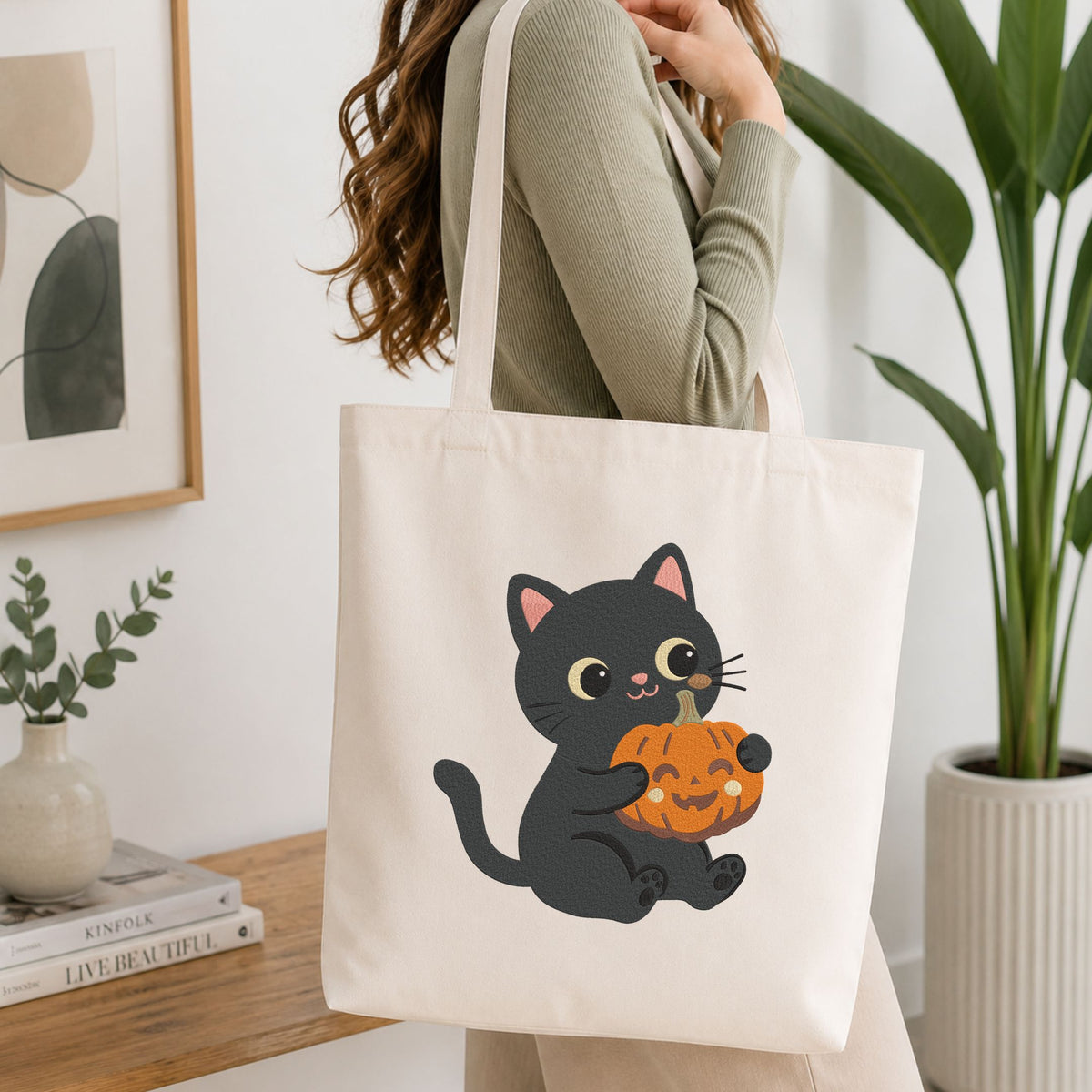  Woman carrying a white natural canvas tote bag featuring a cute black cat holding a mini jack-o-lantern pumpkin machine embroidery design in a bright, modern interior setting   