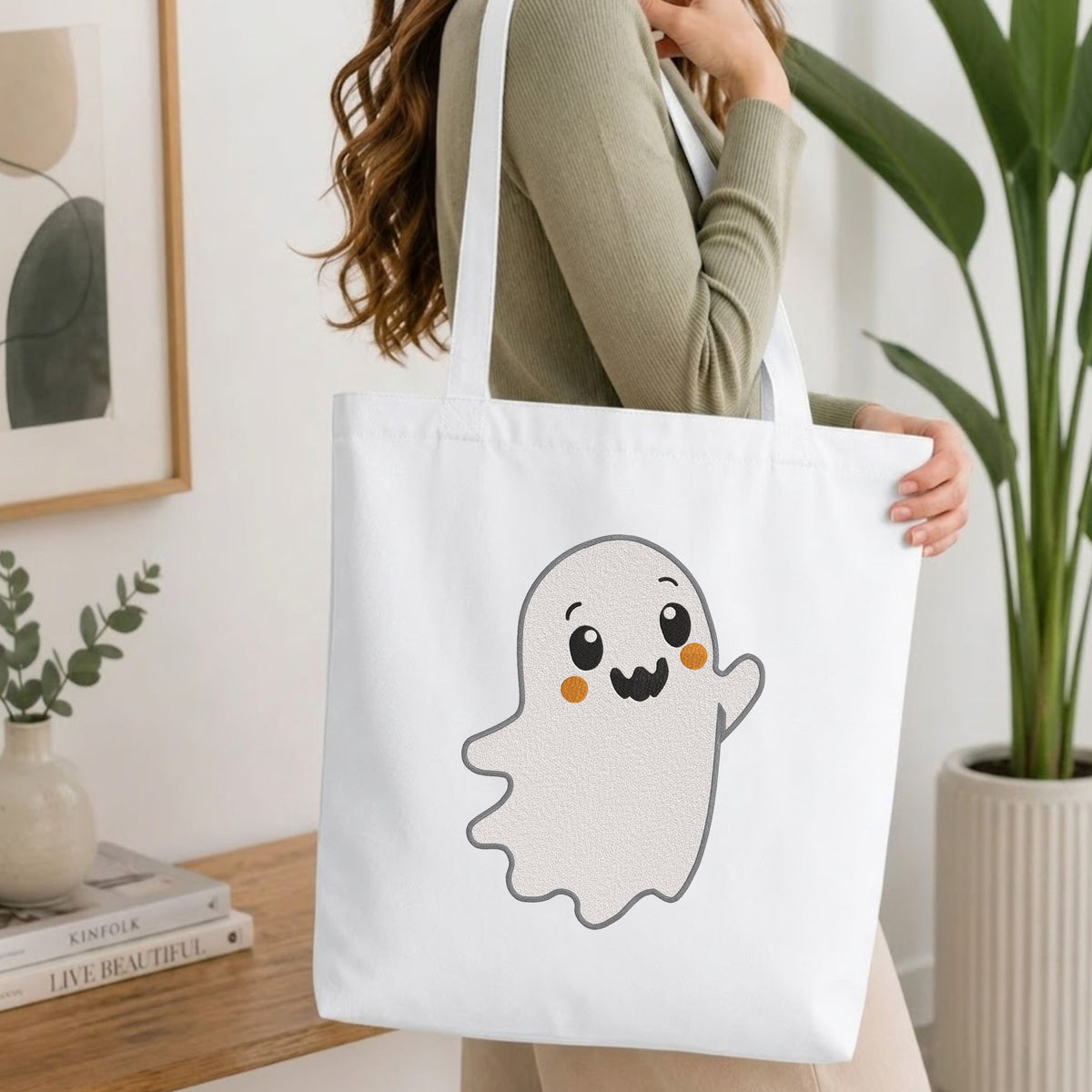 Woman carrying a white natural canvas tote bag featuring a cute waving ghost machine embroidery design in a bright, modern interior setting