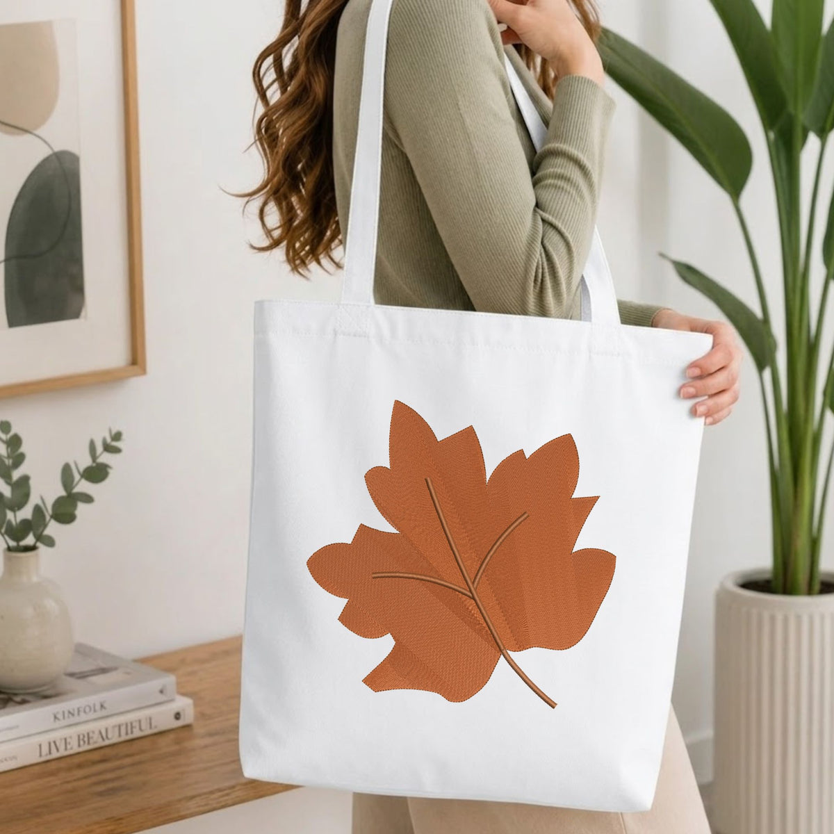 Woman carrying a white natural canvas tote bag featuring an embroidered brown autumn leaf design in a bright, modern interior setting.