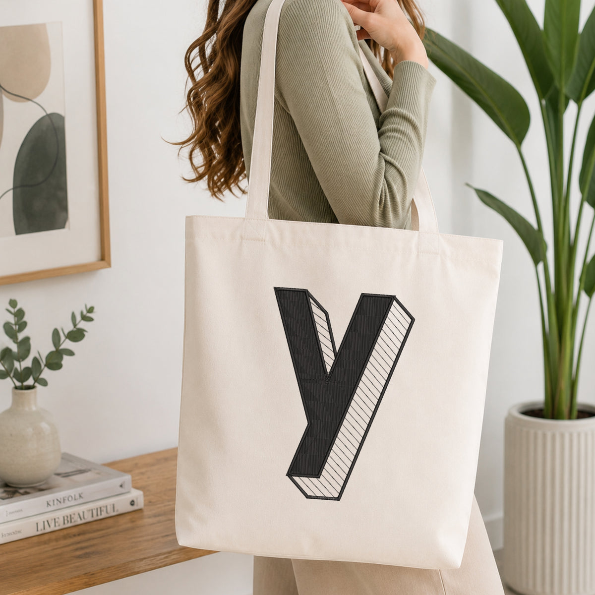 A woman wearing a sage green ribbed long-sleeve top carries a large off-white canvas tote bag with a bold black 3D-style letter 'Y' printed on the front. The scene is set in a bright, modern room with a wooden shelf, framed art, a small plant in a vase, and a larger potted plant in the background.