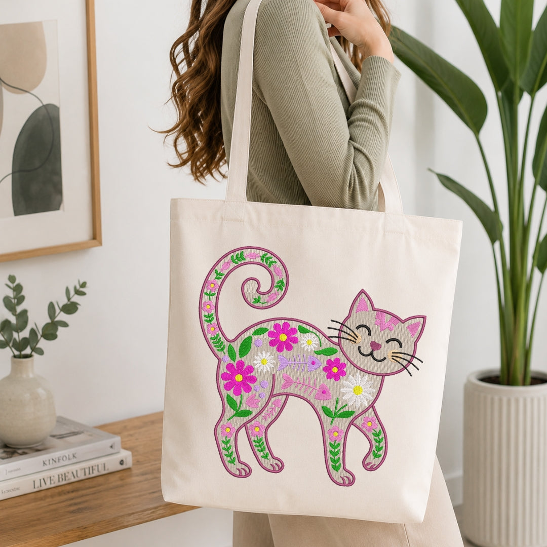 A person wearing a sage green ribbed long-sleeve top carries a cream canvas tote bag decorated with a colorful embroidered floral cat design. The setting is a bright, modern room with a wooden table, books, a small vase, and a large potted plant in the background.