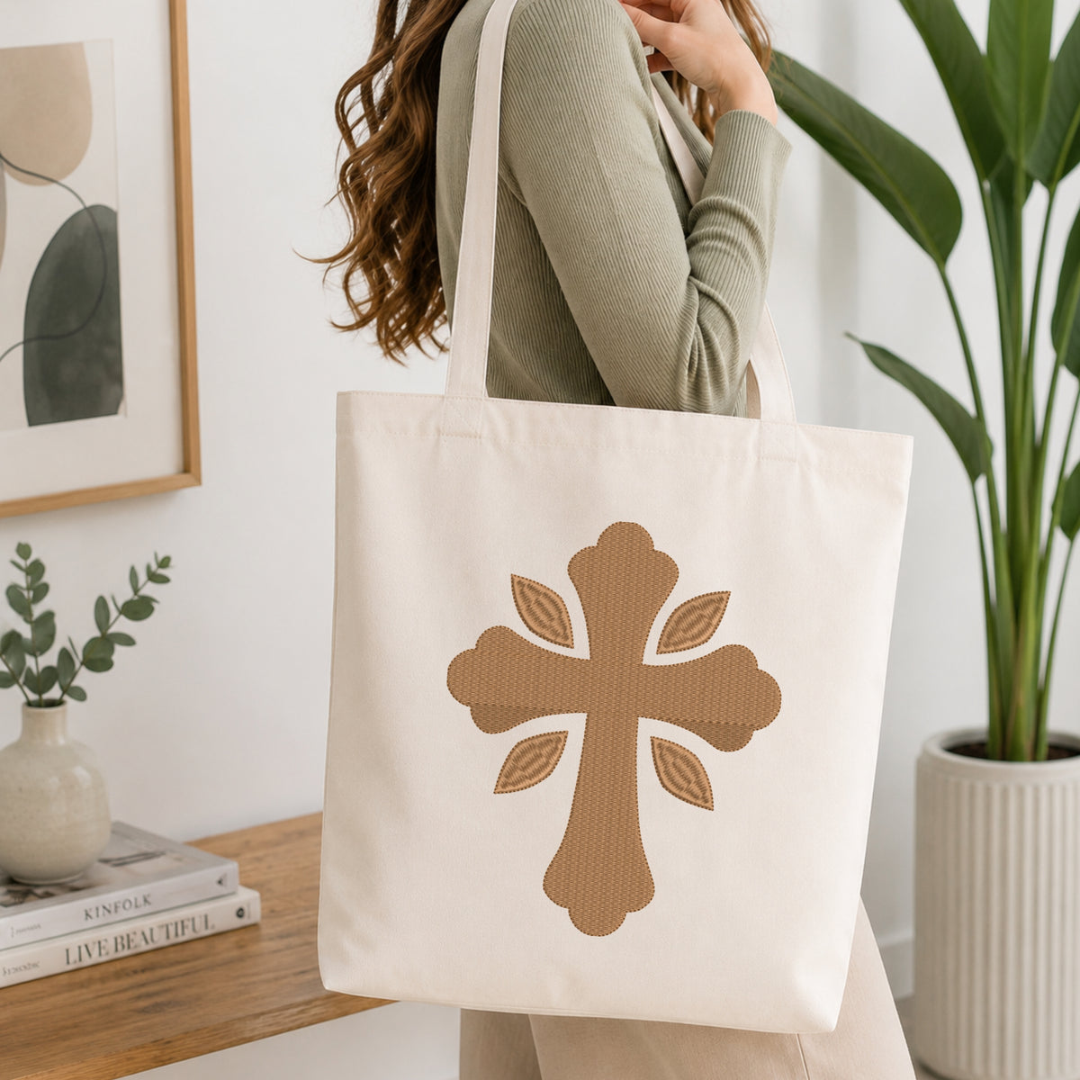 Woman carrying a beige canvas tote bag customized with a gold leaf ornamental cross embroidery design.