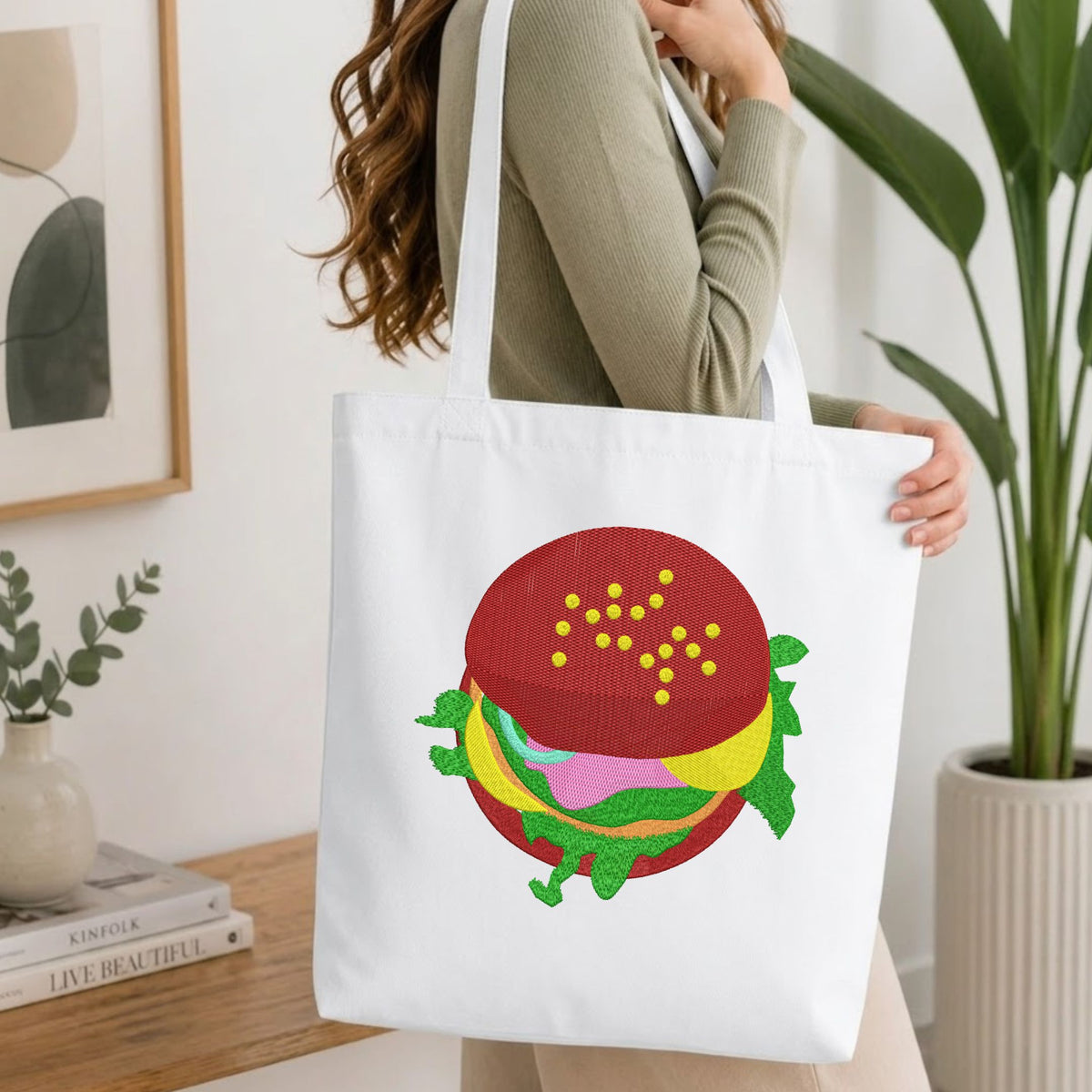 Woman carrying a white natural canvas tote bag featuring an embroidered delicious burger design in a bright, modern interior setting   