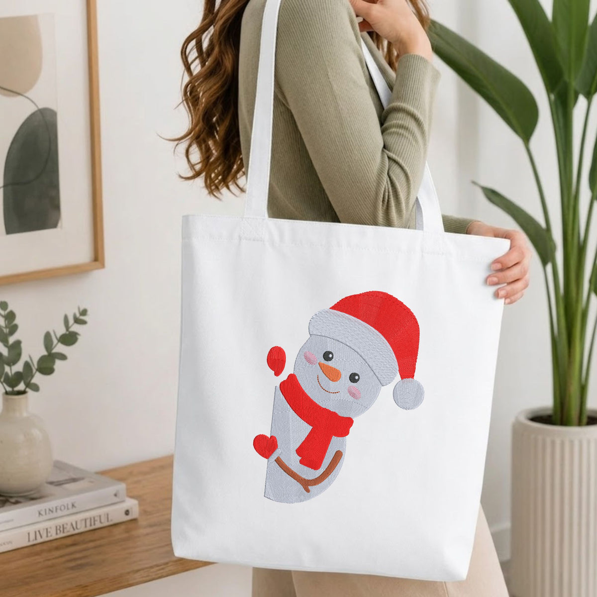  Woman carrying a white natural canvas tote bag featuring an embroidered winter snowman design in a bright, modern interior setting.   