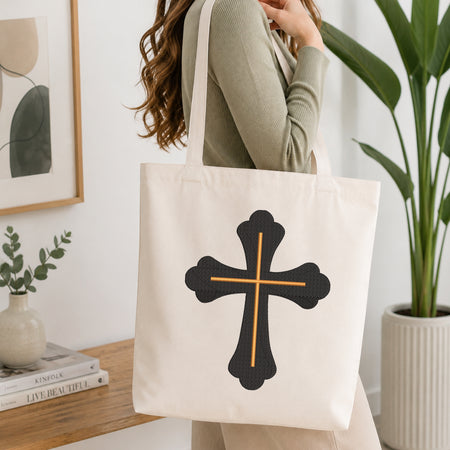 A person wearing a green top holds up a white canvas tote bag printed with the ornate black and gold cross embroidery design, set against a bright, minimalist room background with indoor plants.
