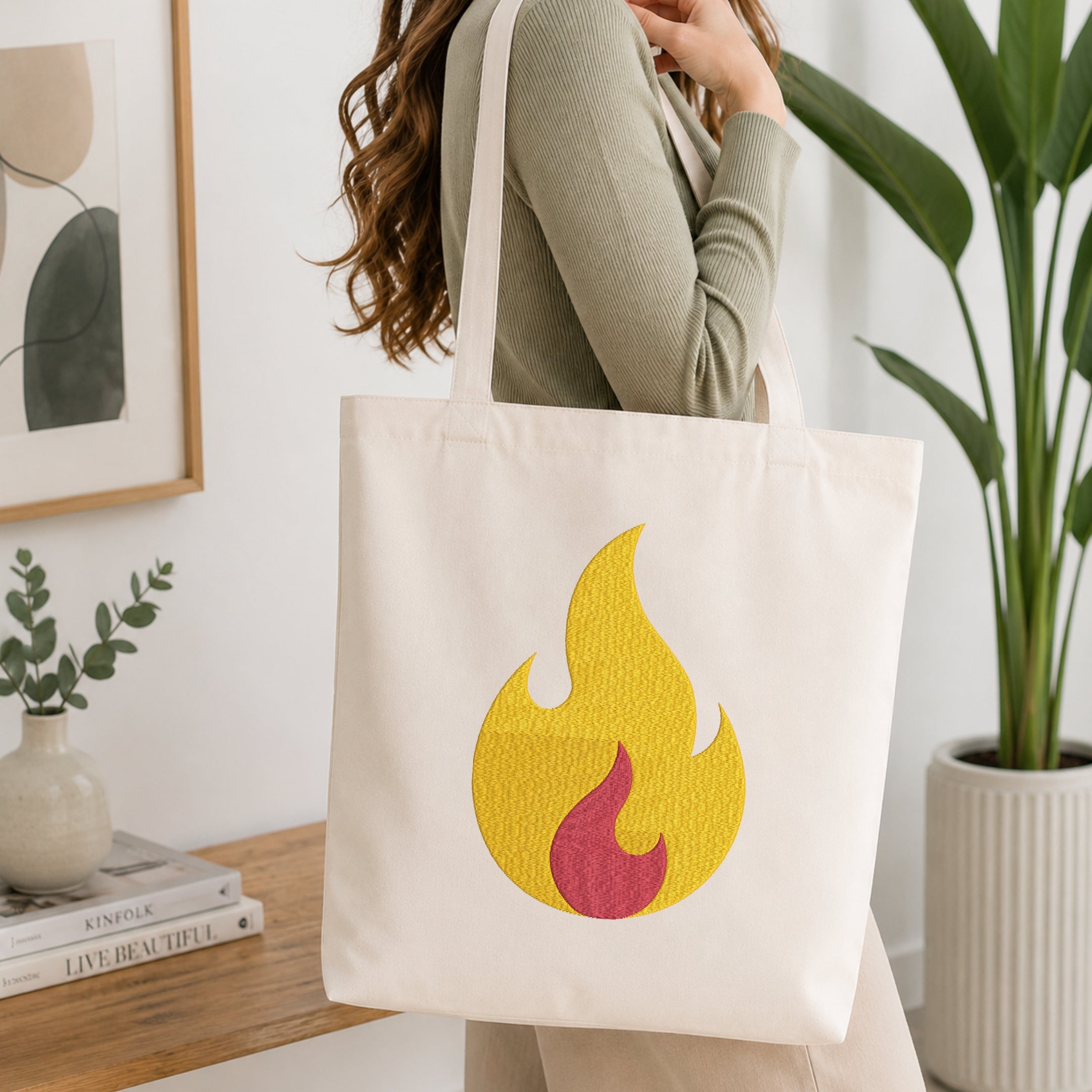 A woman wearing a sage green ribbed sweater carries a cream canvas tote bag with a yellow and red flame graphic on the front, set in a bright, modern room with a wooden table and potted plants.